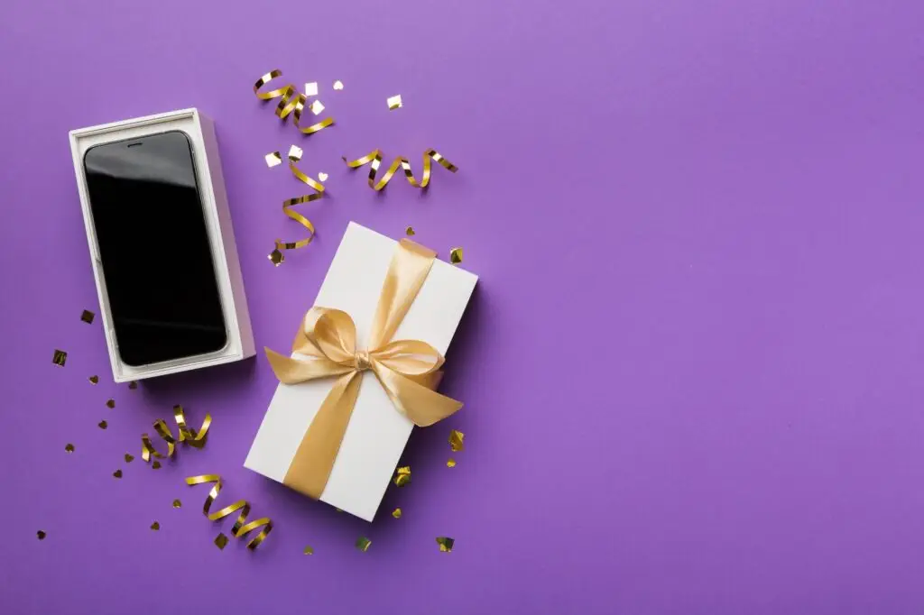 Opened gift box with gold ribbon and smartphone on color background, top view