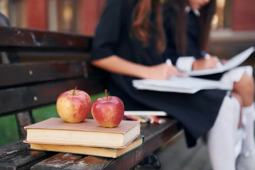 Apples is on the book. Two schoolgirls is outside together near school building